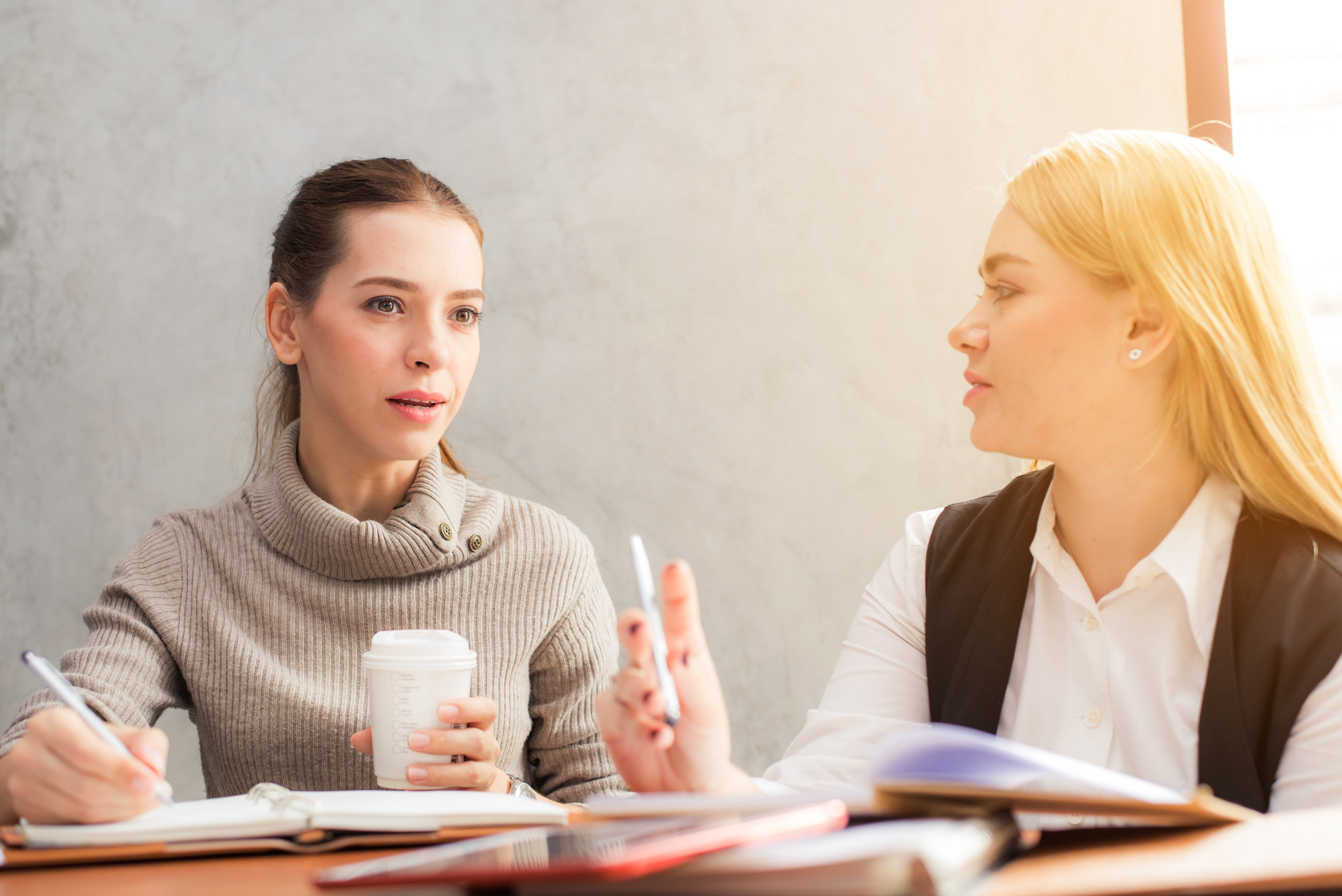 Portrait of two beautiful young girlfriends sitting in modern coffee shop interior and talking with happy smiles. Successful attractive women friends chatting in cafe during coffee break.